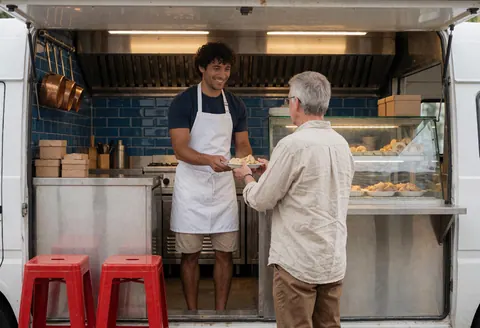Un homme en tablier blanc sert un client à l'intérieur d'un food truck avec des carreaux bleus et des chaises rouges.