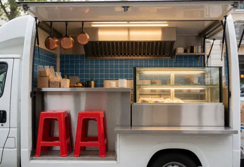 Vue intérieure d'un food truck avec comptoir en acier inoxydable, mur bleu, chaises rouges, vitrine alimentaire et éclairage chaleureux.
