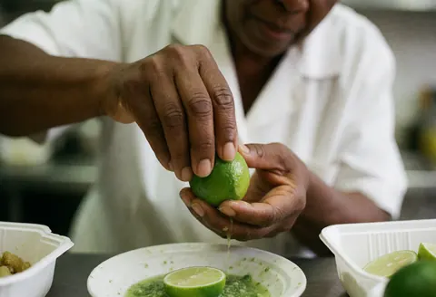 Mains d'un cuisinier pressant un citron frais au-dessus d'une sauce verte dans une assiette, en premier plan avec un arrière-plan flou.