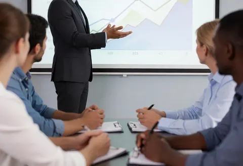 Un professionnel présente des graphiques de croissance sur un écran devant une équipe attentive assise à une table, prenant des notes avec des cahiers et des stylos.