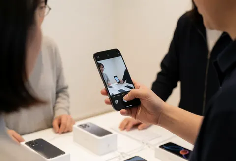 Un vendeur montre un téléphone noir à un client dans une boutique. Des boîtes de smartphones sont sur la table, l'écran affiche l'interface caméra.