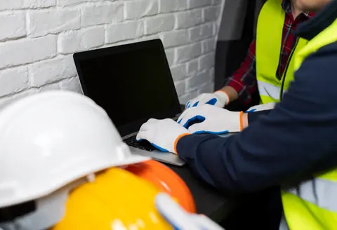 Inspecteur sur chantier portant casque et gilets de sécurité travaillant avec un ordinateur portable devant un mur en briques blanches dans un environnement professionnel réaliste.