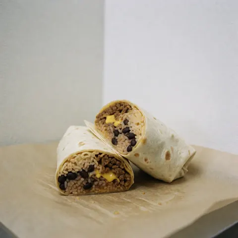 Burrito tranché en deux posé sur du papier sulfurisé, révélant riz, haricots noirs, fromage fondu et viande sur fond blanc.
