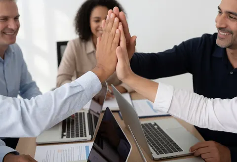 Trois personnes en réunion d'office célèbrent un high five avec ordinateurs portables et documents sur la table dans une ambiance professionnelle et joyeuse.