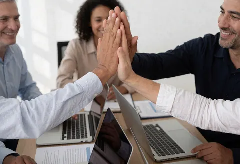 Trois personnes en réunion d'office célèbrent un high five avec ordinateurs portables et documents sur la table dans une ambiance professionnelle et joyeuse.