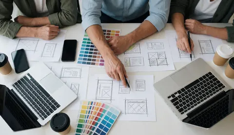 Une équipe de designers travaille sur des croquis de pages web posés sur une table avec des couleurs, ordinateurs, téléphones, café et nuanciers.