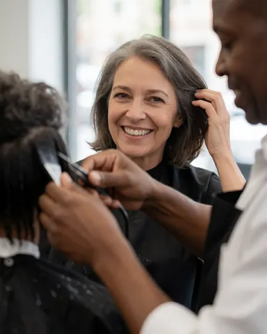 Un coiffeur professionnel utilise des ciseaux et une peigne pour couper les cheveux d'une cliente souriante dans un salon moderne éclairé par la lumière naturelle.