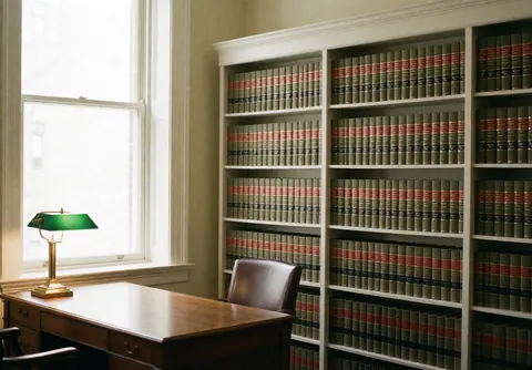 Intérieur d'un cabinet d'avocats avec une bibliothèque remplie de livres de droit, une table en bois, une chaise en cuir et une lampe en bronze, éclairée par la lumière naturelle d'une fenêtre.