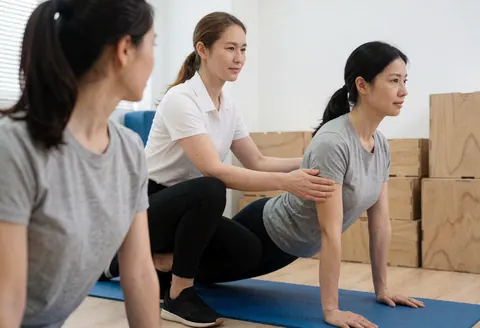 Une instructrice de pilates guide une cliente posée sur un tapis bleu tandis qu'une autre personne observe depuis la gauche dans un espace lumineux.