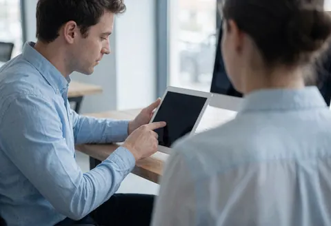 Un agent en chemise bleue explique sur une tablette à un client face à lui dans un bureau avec table en bois.