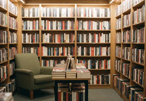 Intérieur d'une librairie avec des étagères en bois remplies de livres, une chaise verte et une table avec des livres empilés, ambiance chaleureuse et accueillante.