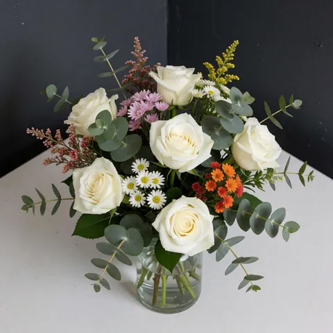 Un centre de table élégant composé de roses blanches et de feuilles d'eucalyptus disposées dans un vase en verre transparent sur une table blanche.