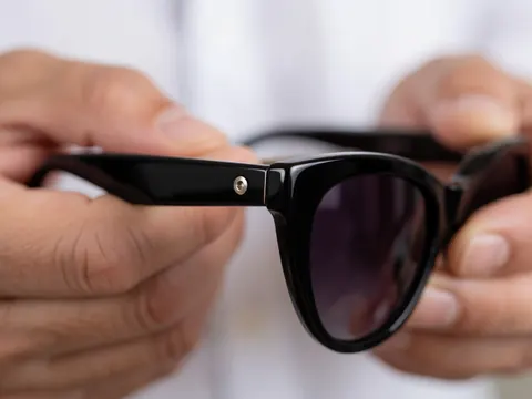Gros plan sur la charnière métallique et les branches en acrylique noir brillant d'une paire de lunettes de style cat eye, tenue par des mains professionnelles sur fond flou.