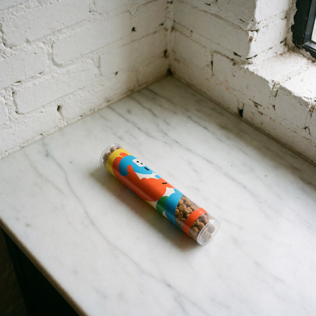 Tube coloré pour friandises canines posé sur un comptoir en marbre blanc avec un mur de briques blanches en arrière-plan et une lumière naturelle.