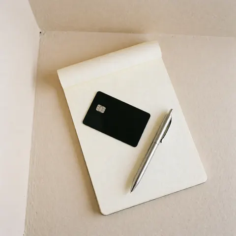 Carte de débit noire et stylo argenté posés sur un carnet en papier crème, scène financière professionnelle avec fond beige.