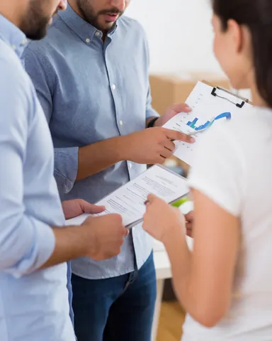 Trois personnes en réunion professionnelle examinant des documents et des graphiques dans un environnement de travail moderne et lumineux.