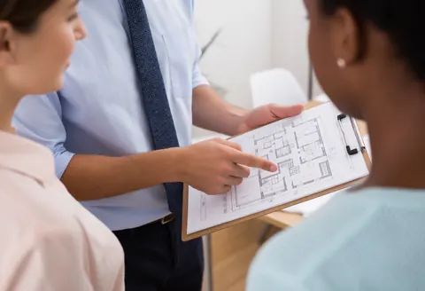 Un conseiller montre des plans architecturaux à deux clients en bureau. Il pointe des détails tandis que les clients observent attentivement, illustrant une réunion professionnelle.