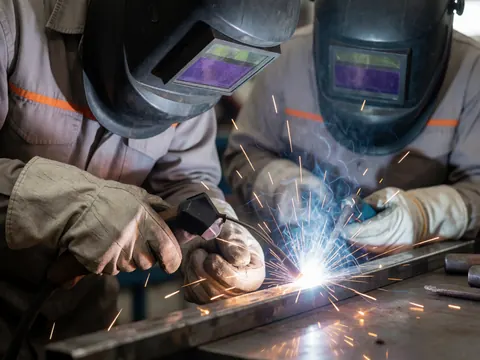 Deux soudeurs équipés de masques et de gants travaillent dans une usine, produisant des étincelles en soudant du métal. Scène industrielle éclairée avec des outils visibles.