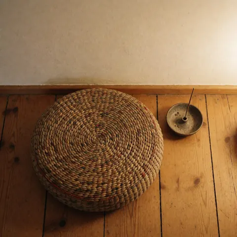 Un coussin de méditation tissé et un porte-encens posés sur un plancher en bois devant un mur blanc, créant une ambiance chaleureuse et relaxante.