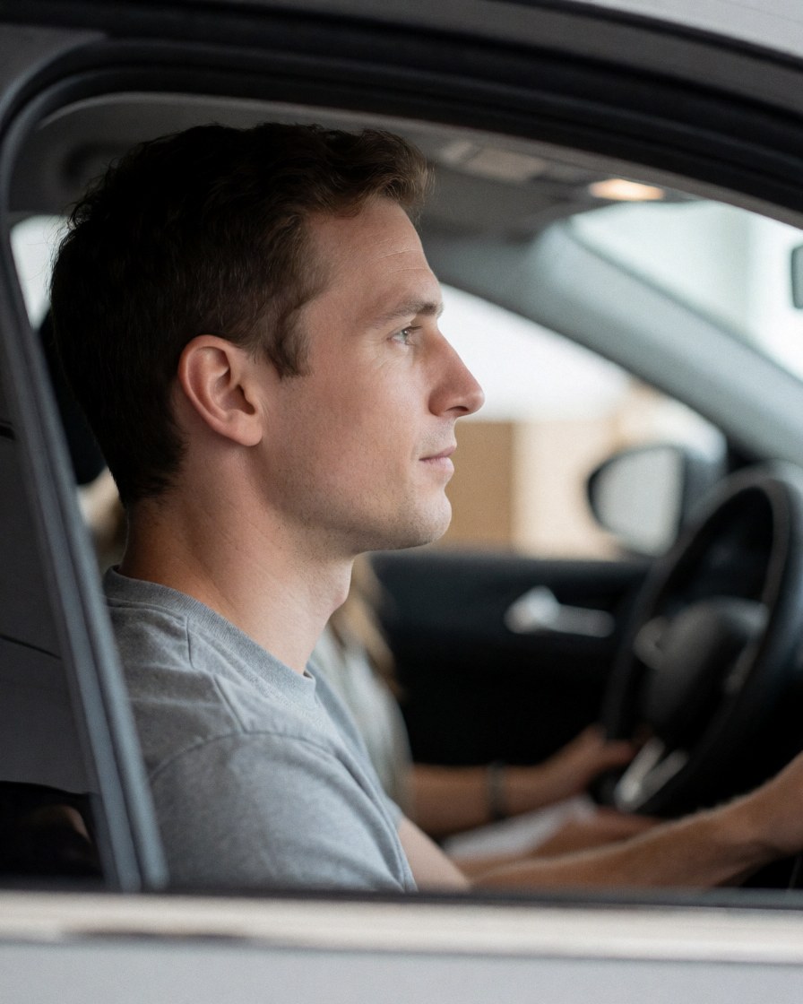 Jeune homme assis au volant d'une voiture, regardant droit devant lui avec concentration, mains sur le volant, intérieur du véhicule visible, arrière-plan flou.