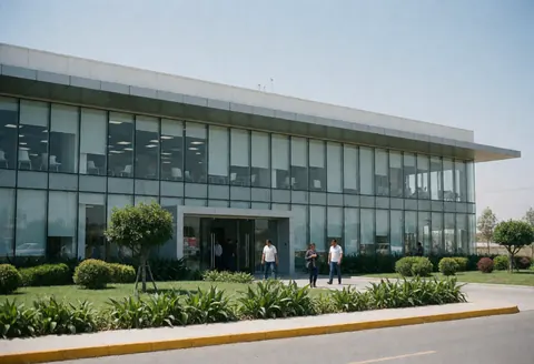 Usine moderne avec façade en verre, jardin verdoyant, personnes marchant à l'extérieur, ciel bleu, entrée principale, architecture propre et professionnelle.