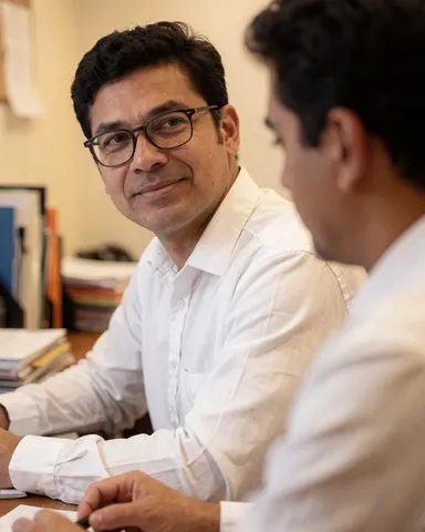 Un parajuriste assis à un bureau en bois avec des documents, des livres et des lunettes, portant une chemise blanche et affichant un sourire professionnel dans un environnement de travail lumineux.