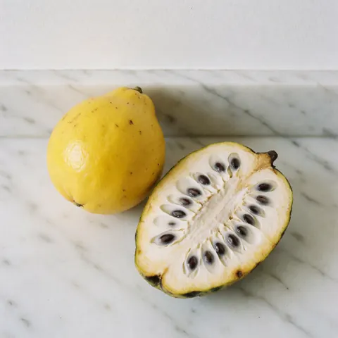 Un lulo entier et un corossol coupé révélant sa pulpe blanche et ses graines noires posés sur un comptoir en marbre blanc éclairé par une lumière naturelle vive.