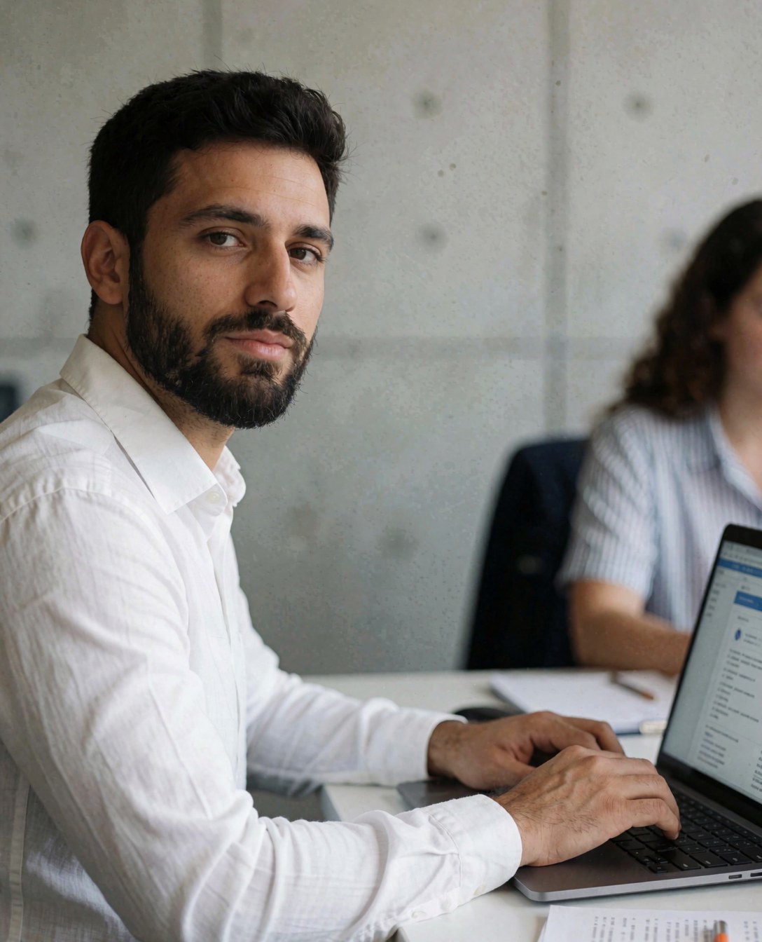 Jeune agent professionnel barbu en chemise blanche travaillant sur un ordinateur portable dans un bureau aux murs de béton, regardant directement l'objectif.