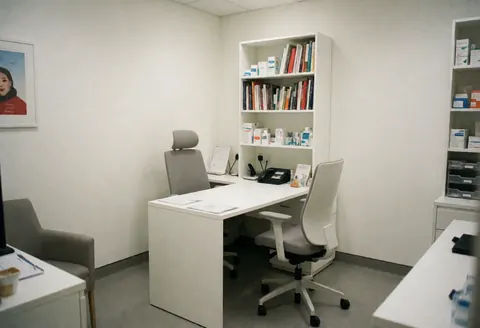 Un consultoire médical moderne avec table blanche, chaises ergonomiques, étagères de livres et médicaments, éclairage neutre et ordre professionnel.