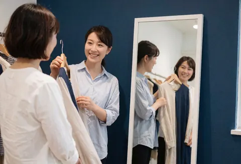 Une styliste montre une blouse à une cliente souriante devant un miroir dans une boutique, sur fond bleu avec lumière naturelle.
