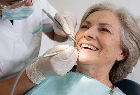 Une hygiéniste dentaire en masque et gants effectue un nettoyage professionnel sur une patiente âgée souriante dans un cabinet lumineux et stérile.