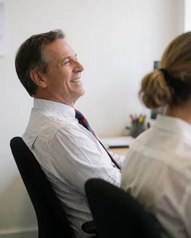 Un homme d'affaires sourit dans un bureau lumineux, assis sur une chaise, avec un collègue dos tourné, ambiance professionnelle.