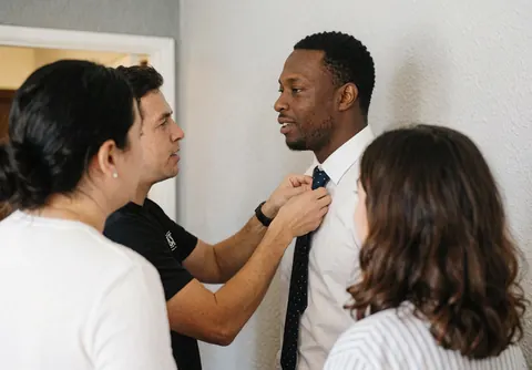 Un organisateur aide le marié à ajuster sa cravate dans un hôtel pendant que deux personnes observent la scène de préparation de la cérémonie.