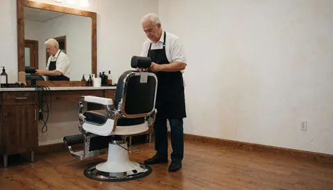 Un barbier expérimenté ajuste un fauteuil de barbier classique dans un salon vide aux murs blancs et au sol en bois, offrant un espace libre à droite pour texte ou logo.