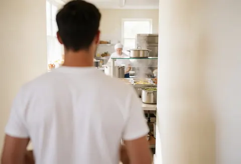 Un volontaire se déplace dans une cuisine communautaire, avec un cuisinier et des casseroles fumantes en arrière-plan, éclairé par la lumière naturelle.