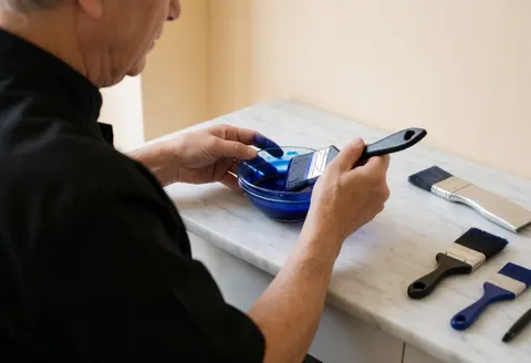 Un barbier âgé prépare une teinture indigo dans un récipient sur une table en marbre blanc, entouré de pinceaux dans un salon aux murs beige et à la lumière naturelle.