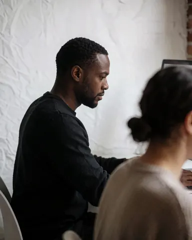 Un homme concentré travaille sur un ordinateur dans un bureau lumineux avec une lumière douce et un mur blanc.