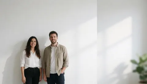 Un couple souriant pose devant un mur blanc avec une lumière naturelle et des ombres douces, idéal pour un fond corporatif ou de présentation.