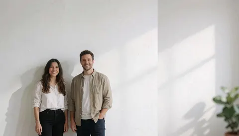 Un couple souriant pose devant un mur blanc avec une lumière naturelle et des ombres douces, idéal pour un fond corporatif ou de présentation.