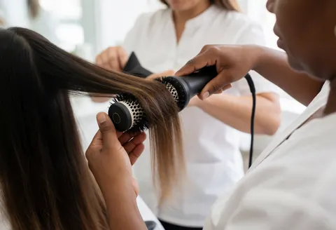 Un coiffeur professionnel utilise un sèche-cheveux et une brosse pour sécher les cheveux longs d'un client dans un salon lumineux et équipé.