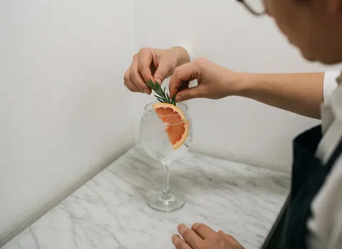 Main posant une tranche de pamplemousse et une branche de romarin sur un gin tonic, sur une surface en marbre blanc, dans un cadre de bar élégant.