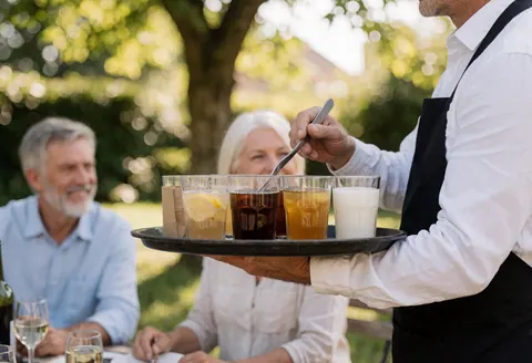 Un serveur transporte un plateau rempli de boissons vers des clients installés dans un jardin en plein air, baigné de lumière naturelle.