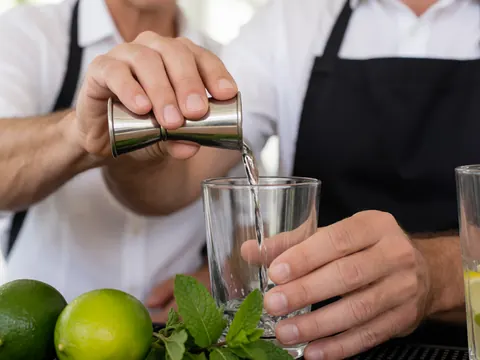 Un bartender prépare un cocktail en utilisant un jigger, des citrons verts et de la menthe fraîche dans une lumière naturelle sur un fond flou.