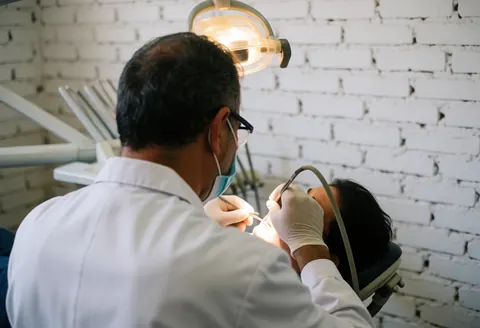 Dentiste en masque et gants examine un patient dans une clinique avec mur en briques blanches et éclairage professionnel.