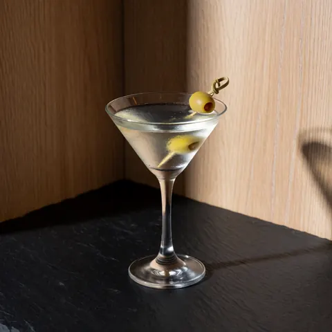 Un martini servi dans une coupe traditionnelle, garni d'une olive piquée sur un cure-dent, posé sur une ardoise sombre devant un mur en bois clair avec un éclairage dramatique.