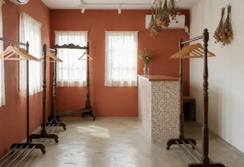 Intérieur d'un magasin avec murs en terre cuite, sol en béton poli, cintres vintage et comptoir en mosaïque. Lumière naturelle par des fenêtres avec rideaux en dentelle.