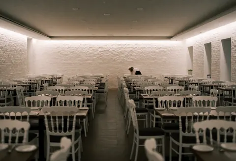 Salle d'événements spacieuse équipée de tables et chaises blanches, murs en briques peintes en blanc et éclairage doux. Une personne travaille au fond.