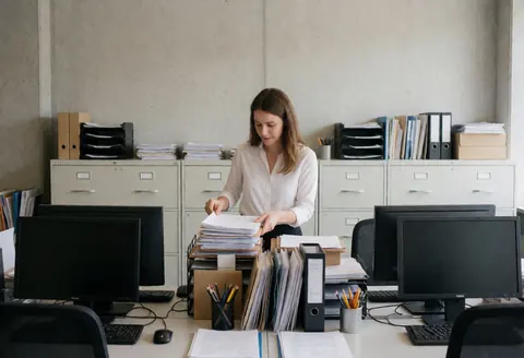 Un manager trie des papiers et des dossiers sur un bureau dans un bureau moderne avec des ordinateurs et des murs en béton.