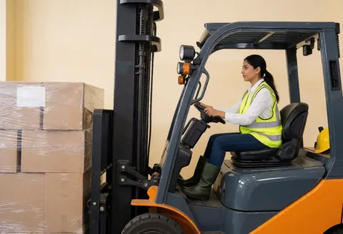 Une femme en gilet réfléchissant et casque de sécurité conduit un chariot élévateur pour déplacer des cartons dans un entrepôt industriel.