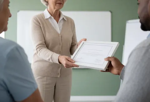 Une instructrice remet un certificat encadré à un étudiant dans une salle de classe moderne. Scène professionnelle et réaliste.